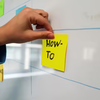 Eine Hand klebt ein gelbes Post-It mit den Worten “How-to” an ein weißes Whiteboard. Im Hintergrund sieht man schemenhaft weitere bunte Post-its.