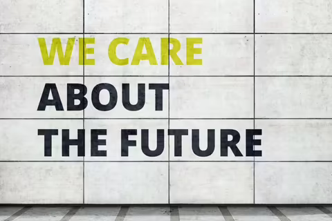 Eine helle Außenwand mit großen, rechteckigen Kacheln. Darauf steht in Großbuchstaben in hellem gelbgrün und dunklem blau in Englisch „we care about the future“. Übersetzung: „Wir kümmern uns um die Zukunft“.