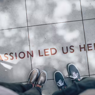 Text “Passion lead us here” geschrieben in Druckbuchstaben auf Bodenkacheln. Davor stehen zwei Personen. Aus der fotografierten Perspektive sieht man nur deren Füße.