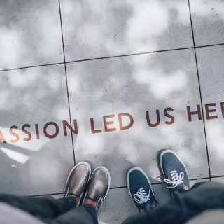 Text “Passion lead us here” geschrieben in Druckbuchstaben auf Bodenkacheln. Davor stehen zwei Personen. Aus der fotografierten Perspektive sieht man nur deren Füße.