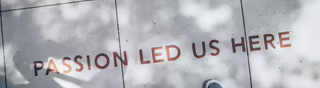 Text “Passion lead us here” geschrieben in Druckbuchstaben auf Bodenkacheln. Davor stehen zwei Personen. Aus der fotografierten Perspektive sieht man nur deren Füße.